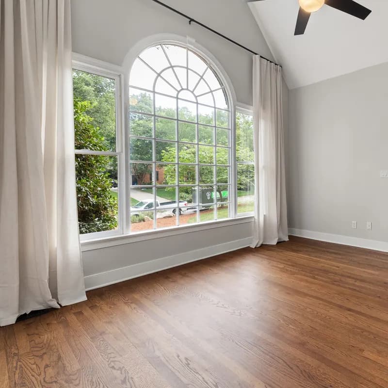 Before: the empty living room of a listing photo, with a vaulted ceiling and fan, a tall arched window with white curtains and a hardwood floor.