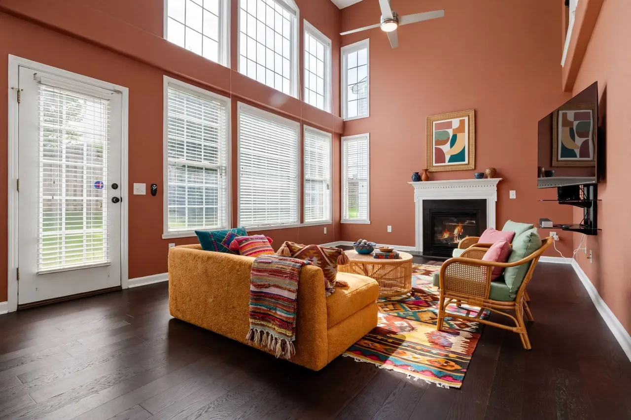 The same living room virtually staged in a Bohemian style: terracotta walls, a mustard boucle sofa with patterned cushions and a striped fringed throw, two rattan armchairs with sage and pink cushions, a round rattan coffee table, a bright kilim rug and an abstract print in a rattan frame above the mantel; the glass door, the windows, the blinds, the TV and the ceiling fan are where they were.