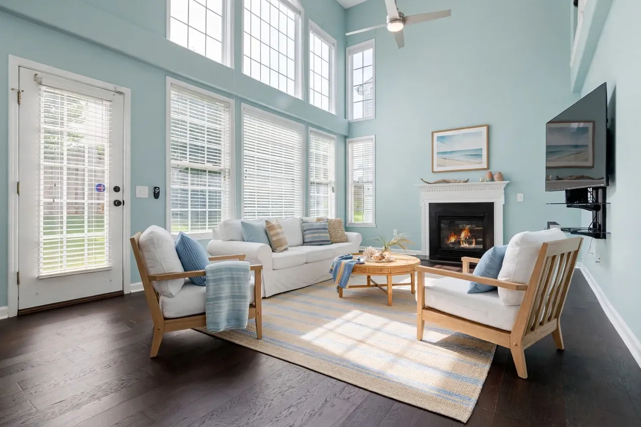The same living room virtually staged in a Coastal style: pale aqua walls, a white slipcovered sofa with blue and striped cushions, two oak armchairs with white cushions, a round rattan coffee table with shells, a blue-and-sand striped rug and a beach watercolour above the mantel; the glass door, the windows, the blinds, the TV and the ceiling fan are where they were.