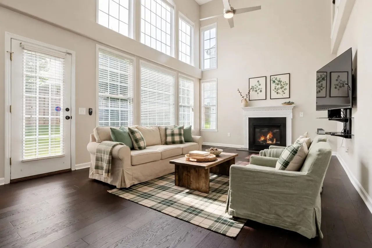 The same living room virtually staged in a Farmhouse style: warm cream walls, a rolled-arm linen sofa with green check cushions and a plaid throw, two slipcovered sage armchairs, a reclaimed-wood coffee table on a green plaid rug and two eucalyptus prints above the mantel; the glass door, the windows, the blinds, the TV and the ceiling fan are where they were.