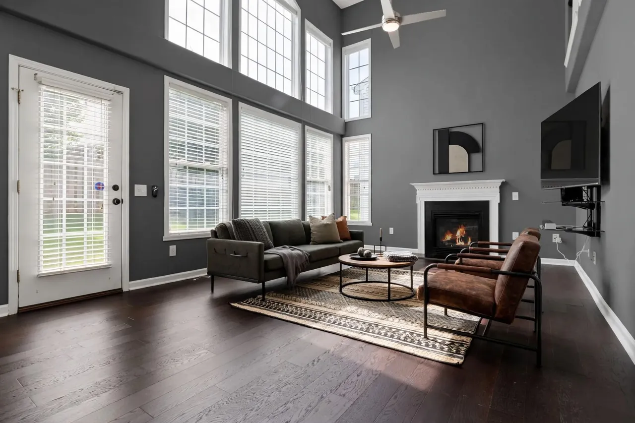 The same living room virtually staged in an Industrial style: slate-grey walls, an olive-grey sofa on black metal legs, two cognac leather sling chairs on black tubular frames, a round wood-and-metal coffee table on a patterned flatweave rug and a large black framed graphic print above the mantel; the glass door, the windows, the blinds, the TV and the ceiling fan are where they were.