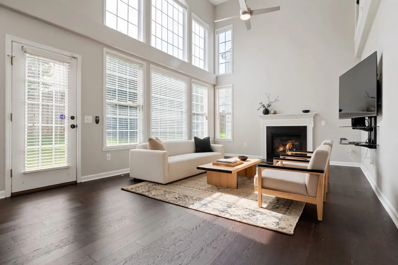 The same living room virtually staged in a Japandi style: soft off-white walls, a low cream sofa with black and beige cushions, two oak armchairs with black arm rails, a low oak coffee table with books and a ceramic bowl, a pale rug and a single branch in a dark vase on the mantel; the glass door, the windows, the blinds, the TV and the ceiling fan are where they were.