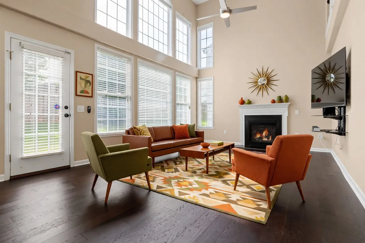 The same living room virtually staged in a Mid-Century style: peach-beige walls, a cognac leather sofa with orange and olive cushions, an olive armchair and a burnt-orange armchair on tapered legs, a teak coffee table on a geometric rug and a sunburst mirror above the mantel; the glass door, the windows, the blinds, the TV and the ceiling fan are where they were.