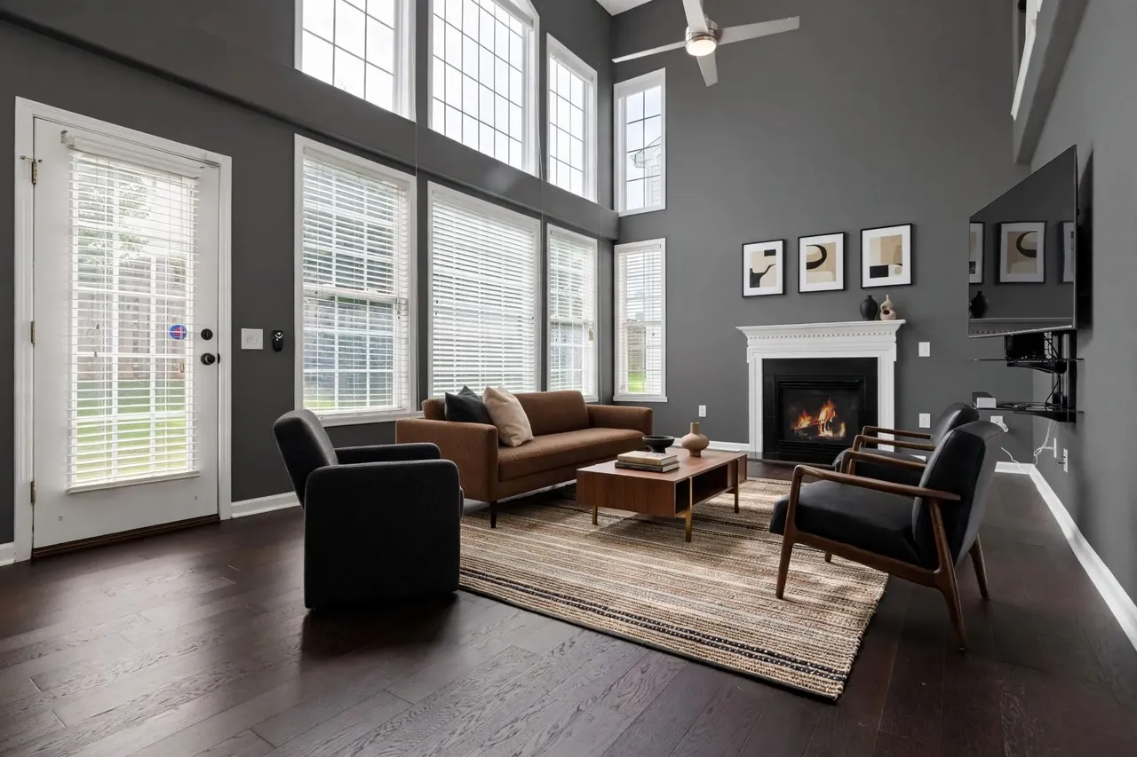 The same living room virtually staged in a Modern style: charcoal walls, a low rust-brown sofa, a black club chair and a black leather lounge chair, a walnut coffee table on a striped jute rug and three abstract prints above the white mantel; the glass door, the windows and their blinds, the TV on its bracket and the ceiling fan are where they were.
