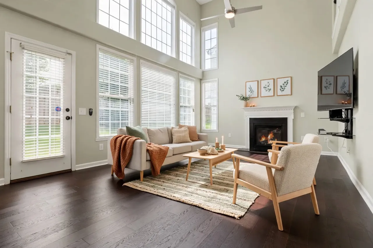 The same living room virtually staged in a Scandinavian style: pale green-grey walls, a cream sofa with a rust knit throw and green cushions, two oak-framed boucle armchairs, a pale oak coffee table with candles, a striped flatweave rug and three botanical prints above the mantel; the glass door, the windows, the blinds, the TV and the ceiling fan are where they were.