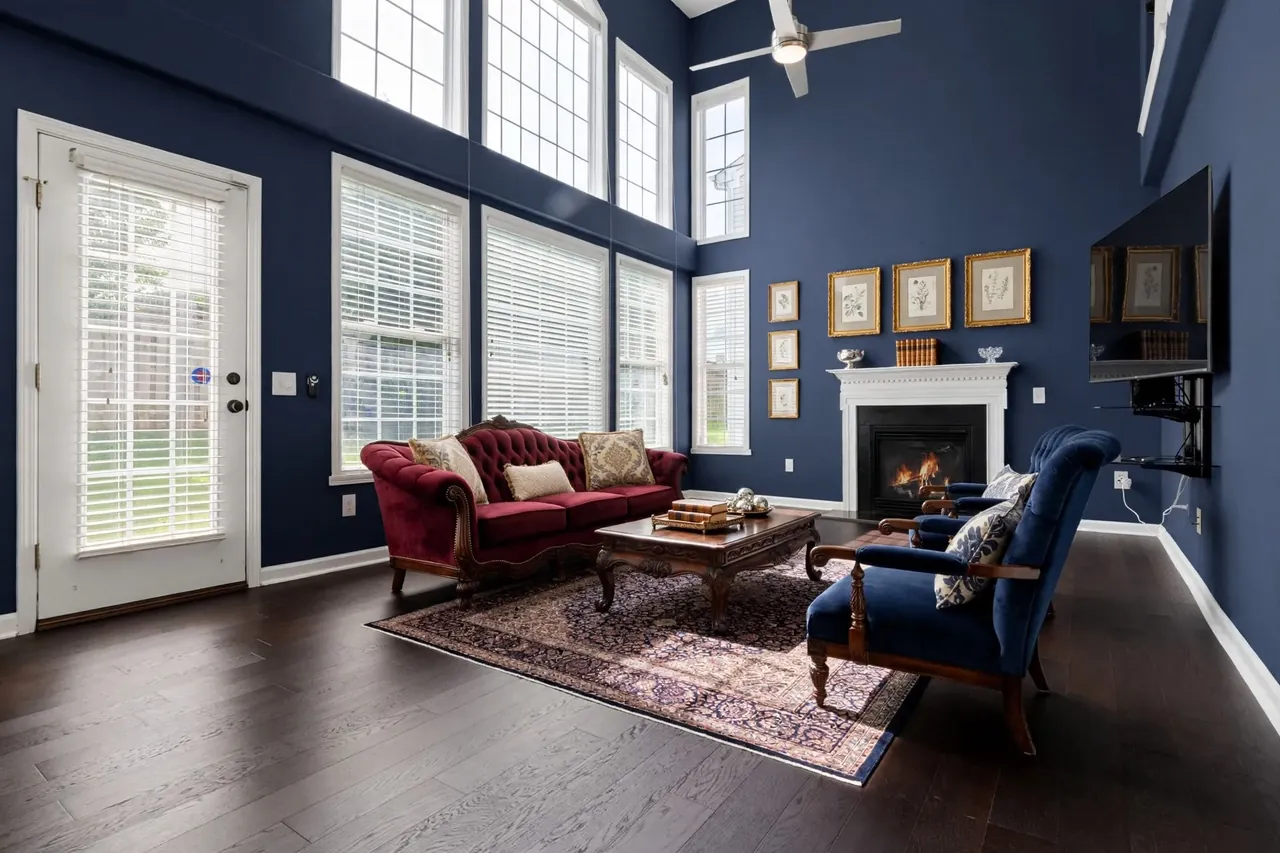 The same living room virtually staged in a Traditional style: navy walls, a carved burgundy velvet camelback sofa, two blue velvet armchairs with carved wood frames, a carved dark wood coffee table on a Persian rug and gilt-framed botanical prints above the mantel; the glass door, the windows, the blinds, the TV and the ceiling fan are where they were.