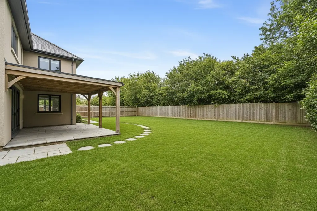 A backyard behind a two-storey house: a covered porch with stone paving, a curved stepping-stone path across a wide lawn, a wooden fence and tall trees behind it.