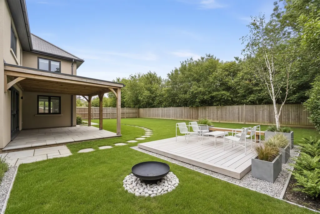 The same backyard in the Scandinavian Minimalist style: a pale wood deck with white chairs, tables and a bench, concrete planters with grasses, a birch tree, ferns in gravel borders and a black fire bowl on white pebbles; the house, covered porch, paving, stepping-stone path, fence and trees are where they were.