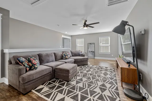 Listing photo of a long family room with a grey sectional sofa, a patterned rug, a TV on a wood console, a floor lamp, three windows and a door at the far end
