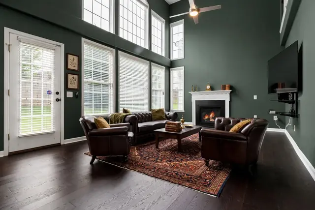 The same room staged in Dark Academia: deep green walls, a brown leather Chesterfield sofa, two leather club armchairs, a Persian rug, a dark wood coffee table with old books and a brass urn, botanical prints and a mantel clock; white mantel, windows, door, TV and fireplace unchanged