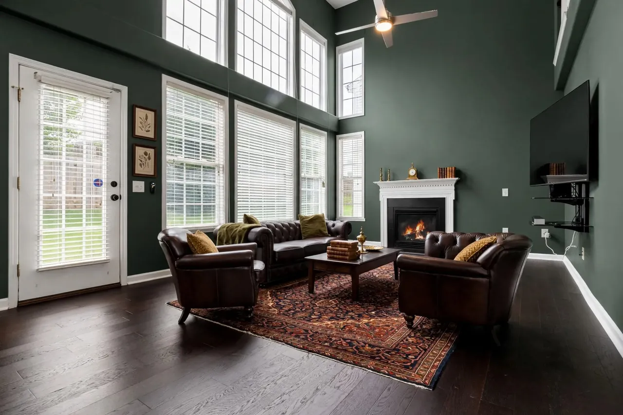 The same room staged in Dark Academia: deep green walls, a brown leather Chesterfield sofa, two leather club armchairs, a Persian rug, a dark wood coffee table with old books and a brass urn, botanical prints and a mantel clock; white mantel, windows, door, TV and fireplace unchanged