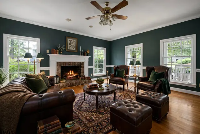 The same living room staged in Dark Academia: deep green walls, a Chesterfield sofa, leather wing chairs, tufted ottomans, a Persian rug and green-shaded brass lamps; brick fireplace, windows, trim and ceiling fan unchanged