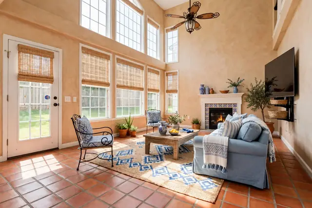 The same room staged in Mediterranean: sand-coloured plaster walls, terracotta floor tiles, bamboo shades, a blue-and-white tiled fireplace surround, a slipcovered sofa, wrought-iron chairs and a jute rug; windows, door and TV unchanged