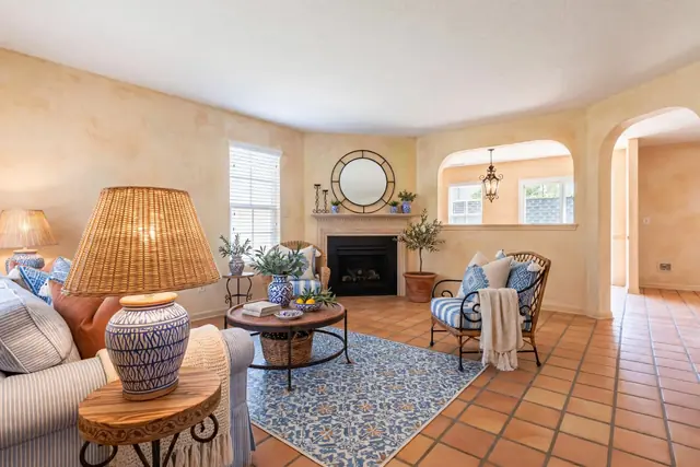 The same room staged in Mediterranean: limewashed walls, a terracotta tile floor, a blue-and-white rug, a striped slipcovered sofa, a rattan chair and ceramic lamps; fireplace, mirror, window, both arches and the pass-through unchanged