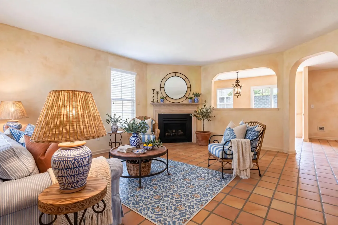The same room staged in Mediterranean: limewashed walls, a terracotta tile floor, a blue-and-white rug, a striped slipcovered sofa, a rattan chair and ceramic lamps; fireplace, mirror, window, both arches and the pass-through unchanged