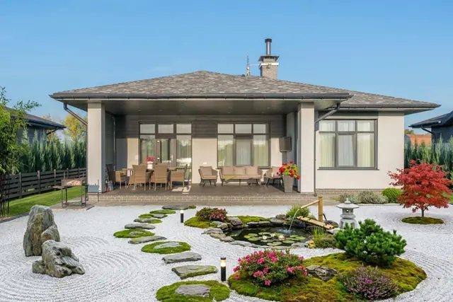 The same backyard restyled as a Japanese Zen garden: raked white gravel in place of the lawn, moss islands with weathered rocks, stepping stones, a koi pond with water lilies and a bamboo fountain, a stone lantern, pink azaleas, a dwarf pine and a red maple in the far right corner; house, windows, roof, covered terrace, furniture and barbecue unchanged