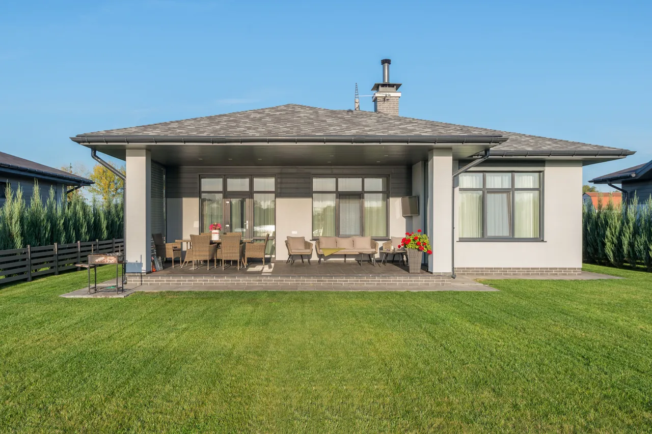 Listing photo of the back of a single-storey house: a covered terrace with wicker dining and lounge furniture, a barbecue, a large green lawn and a hedge along the fence.