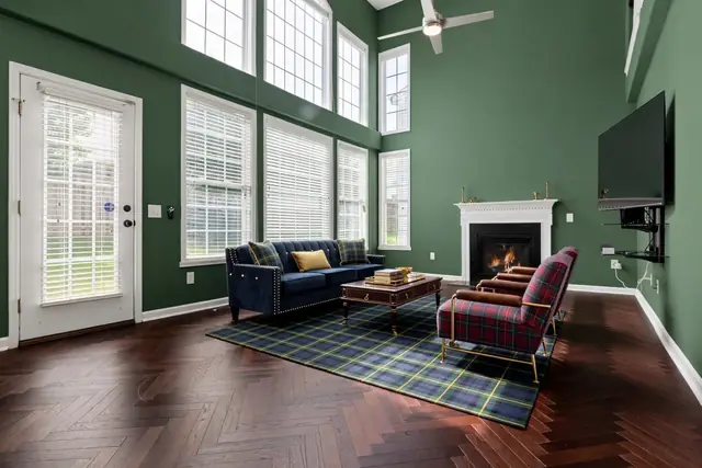 The same room staged in Preppy Ivy League: hunter green walls, a herringbone wood floor, a navy velvet sofa, red tartan armchairs on brass frames, a navy-and-green tartan rug, a mahogany coffee table and brass candlesticks; windows, door, fireplace and TV unchanged
