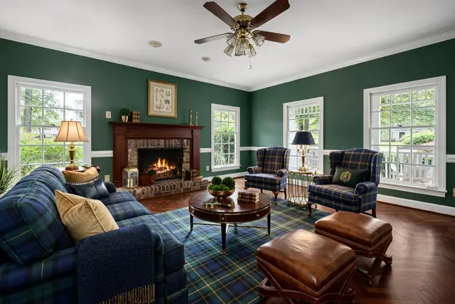 The same living room staged in Preppy Ivy League: hunter green walls, a herringbone floor, a mahogany mantel, a tartan sofa and wing chairs, leather ottomans and brass lamps; brick firebox, windows, trim and ceiling fan unchanged