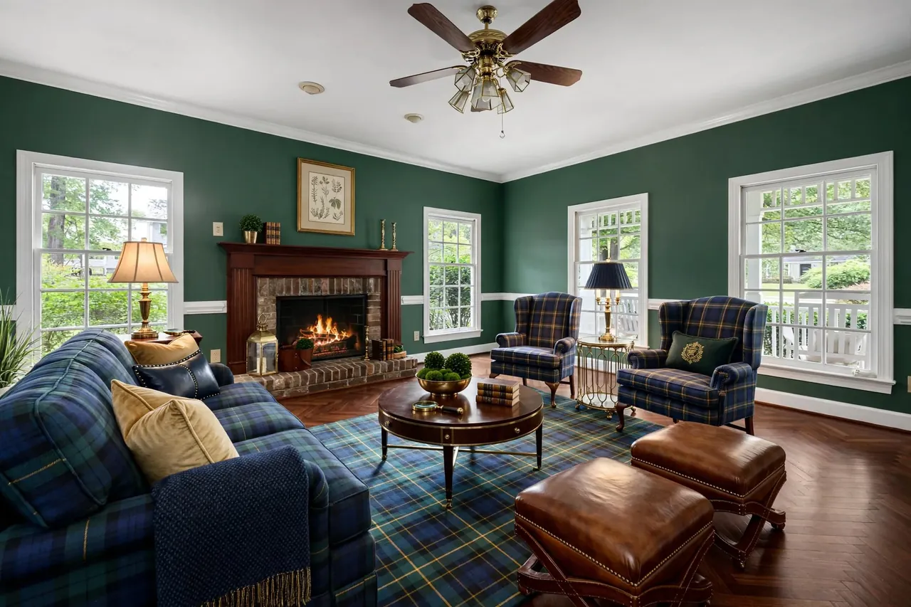 The same living room staged in Preppy Ivy League: hunter green walls, a herringbone floor, a mahogany mantel, a tartan sofa and wing chairs, leather ottomans and brass lamps; brick firebox, windows, trim and ceiling fan unchanged